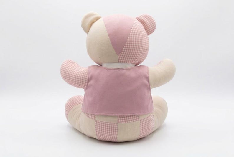 May include: A stuffed teddy bear featuring a patchwork design of cream-colored fabric and pink gingham fabric. The bear is wearing a solid pink vest over its torso and has a white ribbon collar around its neck, viewed from the back.