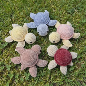 May include: Five plush crochet turtles in various colors including light blue, pale yellow, soft pink, dusty rose, and deep maroon. Each turtle features a rounded shell, four flippers, and a small head with two black bead eyes.