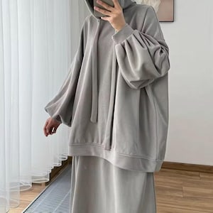 May include: A light gray hoodie and matching skirt set. The hoodie features a drawstring hood and long sleeves with a voluminous, gathered design. The skirt is a simple, flowing style. The outfit is made of a soft, comfortable fabric.