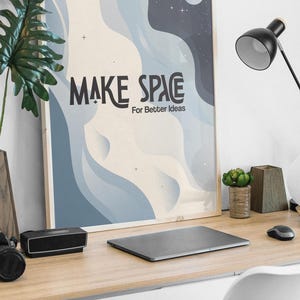 Puede incluir: Una impresión artística enmarcada con las palabras "MAKE SPACE For Better Ideas" en texto negro. La obra de arte presenta un diseño abstracto azul y blanco con una luna y estrellas. La impresión se muestra en un escritorio de madera con una computadora portátil, una lámpara y elementos decorativos.