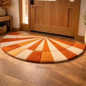 Puede incluir: Una alfombra semicircular con un diseño de rayos de sol en tonos naranja, crema y blanco. La alfombra está colocada sobre un suelo de madera cerca de una puerta de madera. Mide aproximadamente 91 cm de ancho y 183 cm de largo.