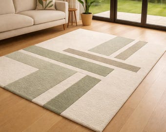 May include: A rectangular area rug with a geometric design. The rug features a cream-colored background with rectangular and linear accents in shades of green and tan. The rug is placed on a hardwood floor.