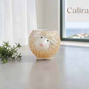 Può includere: Un vaso di ceramica a forma di pecora, color crema con viso bianco, naso rosa e occhi neri. La parola "Calira" è visibile sullo sfondo. Progettato per piccole piante.