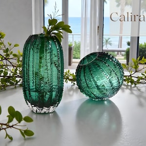 Puede incluir: Dos jarrones decorativos de cristal verde turquesa. Uno es alto y cilíndrico, el otro esférico. Los jarrones tienen un diseño texturizado y acanalado. La palabra "Calira" es visible.