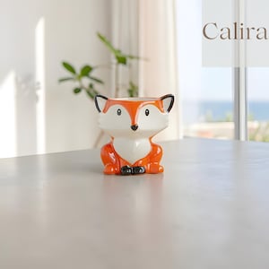 May include: A ceramic fox-shaped planter in orange, white, and black. The planter has a detailed fox face and body, with a hollowed-out top for plants. The word "Calira" is in the upper right corner. The planter is on a light grey surface.