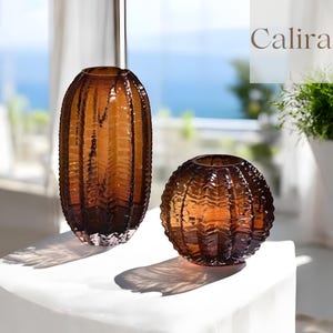 Puede incluir: Dos jarrones de cristal color ámbar con diseños acanalados texturizados, verticales y horizontales. Uno es de forma ovalada, el otro es redondo. Los jarrones están sobre una superficie blanca con un fondo borroso del océano y vegetación. La palabra "Calira" está en la esquina superior derecha.