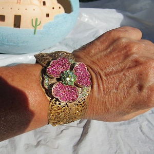 May include: A gold-tone bracelet with a pink and green flower design. The flower is made of rhinestones and is the focal point of the bracelet.