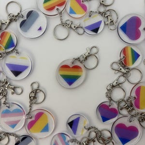 May include: A collection of clear acrylic keychains with various LGBTQ+ pride flag designs. The keychains feature heart and circle shapes with rainbow, transgender, bisexual, non-binary, and pansexual flag patterns. Each keychain has a silver-colored clasp and ring.