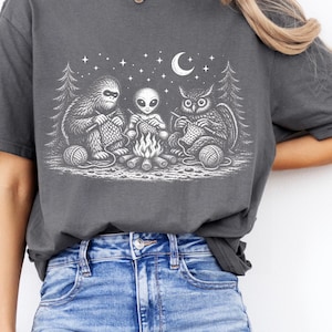May include: Dark gray t-shirt featuring a whimsical illustration of a Bigfoot, an alien, and an owl knitting around a campfire under a starry night sky. The design includes a crescent moon and pine trees, with balls of yarn. A fun, unique graphic tee.