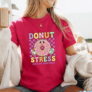 Può includere: Una maglietta magenta con un motivo a ciambella a cartoni animati. La ciambella ha una faccia sorridente e tiene in mano una matita e un rametto di foglie verdi. Il testo sulla maglietta recita "DONUT STRESS JUST DO YOUR BEST".