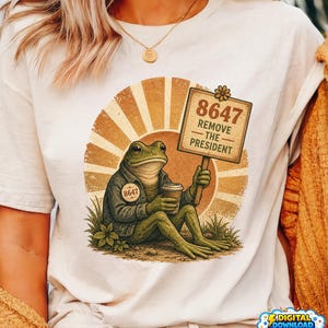 Peut inclure: T-shirt crème avec un dessin d'une grenouille en veste, tenant un gobelet de café. La grenouille tient une pancarte avec l'inscription "8647 Remove the President". Le design comprend un fond rétro en forme de soleil.