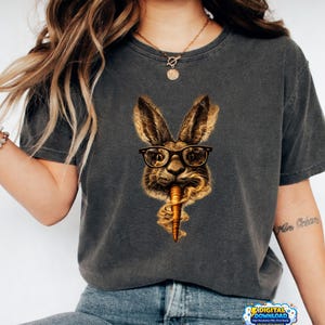 May include: Dark gray t-shirt featuring a rabbit wearing glasses and holding a carrot. The rabbit is brown and tan with black-rimmed glasses. The carrot is orange. The shirt is a short-sleeved crew neck.