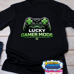 Peut inclure: T-shirt noir avec un motif de manette de jeu verte et noire, des trèfles à quatre feuilles et le texte "LUCKY GAMER MODE". Le motif comprend un symbole de bouton d'alimentation. Le t-shirt est présenté sur une surface en bois clair.