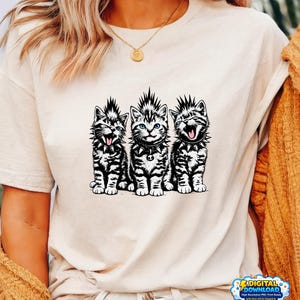 Peut inclure: T-shirt crème avec un motif de trois chats punk rock. Les chats ont des colliers à pointes et des coiffures mohawk. Les chats ont une fourrure rayée noire et blanche et sont assis côte à côte. Les chats ont les yeux bleus et sourient.