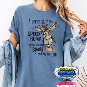 Peut inclure: Un t-shirt bleu avec le texte "I seriously need a SPEED BUMP between my BRAIN and my MOUTH" et un visage de dessin animé d'âne. Le t-shirt a des manches retroussées.