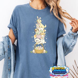 May include: A blue t-shirt features a stack of cartoon dogs, some wearing Easter bunny ears and outfits. A basket of Easter eggs sits at the bottom. The shirt also has the text "DIGITAL DOWNLOAD" and the brand name "Rymel".