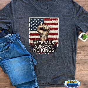 Peut inclure: T-shirt gris foncé à col en V avec un drapeau américain et un poing serré. Le texte "VETERANS SUPPORT NO KINGS" est en dessous. Un jean bleu plié est posé à côté du t-shirt sur une surface en bois.