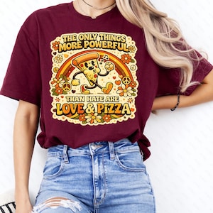 Peut inclure: T-shirt bordeaux avec un motif coloré représentant une part de pizza, un arc-en-ciel et des fleurs. Le texte dit : "THE ONLY THINGS MORE POWERFUL THAN HATE ARE LOVE & PIZZA". Le t-shirt est porté avec un jean bleu.