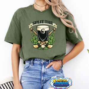 Peut inclure: T-shirt vert olive avec l'inscription "SPLIT THE G" au-dessus d'un dessin d'une pinte de stout, de crosses croisées et de trèfles. La personne porte un jean bleu.
