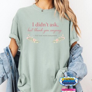 Peut inclure: T-shirt vert sauge avec le texte "I didn't ask, but thank you anyway." en script rose. Le t-shirt présente des motifs floraux et la phrase "first time moms everywhere." Une veste en jean est drapée sur les épaules.