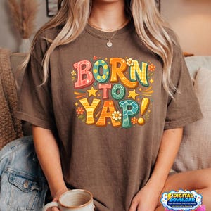 Peut inclure: T-shirt marron avec l'inscription "BORN TO YAP!" dans une typographie colorée et rétro, ornée d'étoiles et de motifs floraux. Le t-shirt est de style décontracté.