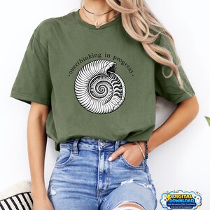 Peut inclure: T-shirt vert olive avec un motif de coquillage nautile blanc et le texte ".overthinking in progress.". Le t-shirt est porté avec un jean bleu. Le motif est un téléchargement numérique.