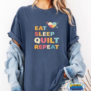 Peut inclure: T-shirt bleu marine avec l'inscription "EAT SLEEP QUILT REPEAT" en lettres colorées. Un motif en forme de cœur avec un design patchwork est au-dessus du texte. Le t-shirt est de style décontracté.