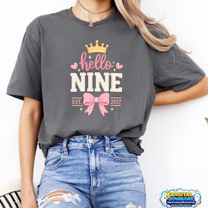 Peut inclure: T-shirt gris foncé avec un motif « hello NINE » rose et blanc, une couronne dorée et un nœud rose. Le texte « EST. 2017 » est sous le nœud. Le t-shirt est de style décontracté.