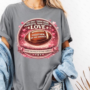 Peut inclure: T-shirt gris avec un motif de ballon de football. Le motif comprend le texte "The only thing more powerful than hate is love" et "Together, we are America". Une bannière en bas indique "From the field to the family".