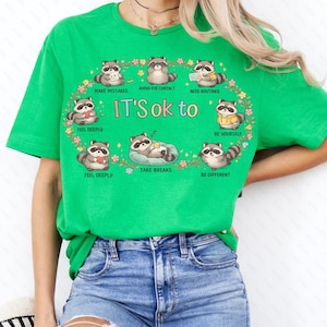 Peut inclure: T-shirt vert vif avec un motif circulaire représentant des ratons laveurs et le texte "IT'S ok to." Le motif comprend des phrases telles que "Faire des erreurs", "Éviter le contact visuel" et "Faire des pauses". Le t-shirt est en matière souple.