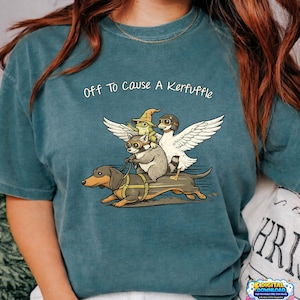 Pode incluir: Camiseta azul-petróleo com a frase "Off To Cause A Kerfuffle" e uma ilustração fantasiosa. O desenho mostra um cachorro salsicha puxando uma carroça com um guaxinim, um sapo e um pato com óculos e asas.