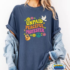Peut inclure: T-shirt bleu marine avec l'inscription "UNPAID PEACEFUL PROTESTER" dans un style rétro coloré. Le design comprend une guitare, un symbole de paix, des fleurs et une colombe. Le t-shirt est de coupe décontractée.