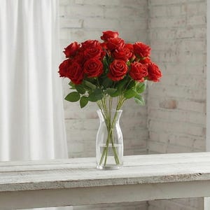 Puede incluir: Un jarrón de cristal transparente lleno de un ramo de rosas rojas. Las rosas tienen hojas verdes y están dispuestas en un jarrón claro y delgado. El jarrón está sobre una mesa de madera blanca, con una pared de ladrillos blancos al fondo.