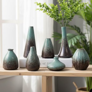 Puede incluir: Una colección de jarrones de cerámica de varias formas y tamaños, con un esmalte de dos tonos turquesa y marrón. Algunos jarrones contienen tallos verdes y frondosos, dispuestos sobre una superficie de madera clara. El fondo incluye una ventana y vegetación.
