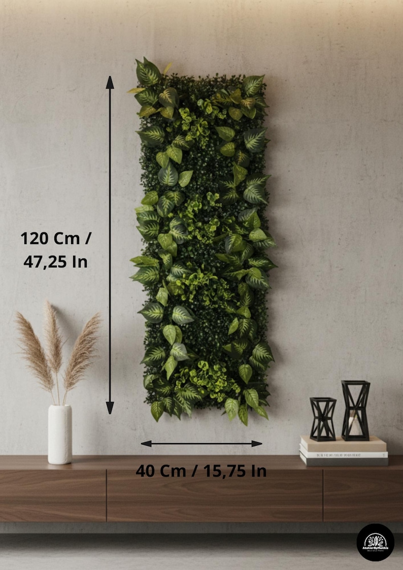 May include: A rectangular vertical artificial plant wall, 47.25 inches tall and 15.75 inches wide, with various shades of green foliage. The wall is mounted on a light gray wall. A brown wooden shelf below holds a white vase with pampas grass and black candle holders.