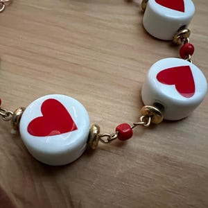 May include: A necklace with white, circular beads, each displaying a red heart. The beads are linked by gold-coloured chain and small red beads. The necklace is set against a wooden background.