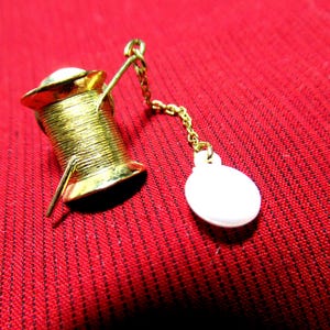 May include: A gold-colored spool of thread pin with a white charm attached by a gold chain. The pin is set against a red background.