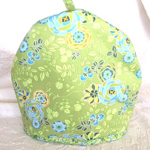 May include: A green tea cozy with a floral pattern featuring blue, yellow, and white flowers and green leaves. The cozy has a loop at the top and a decorative trim at the bottom. The background is a white, textured surface.