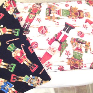 May include: Two Christmas-themed fabric pieces. One is black with a pattern of colorful nutcrackers, candy canes, and stars. The other is white with a similar pattern, including Santa Claus figures and ornaments. The fabrics are likely for holiday crafts.
