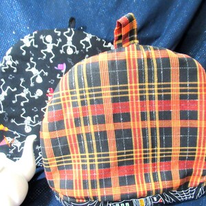 May include: Two tea cozies. One is black with white skeleton figures and musical notes. The other is a plaid pattern in red, yellow, and black. Both have a loop at the top. The bottom of the plaid cozy has a black and white spiderweb design.