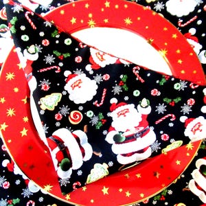 May include: A festive Christmas table setting featuring a red plate with gold stars and a black napkin. The napkin is decorated with Santa Claus, candy canes, snowflakes, and other holiday-themed elements. The overall theme is a cheerful holiday dinner.