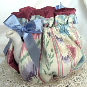 May include: A patterned teapot cosy with a light blue bow. The cosy is made of fabric with a cream background and vertical stripes in pink, green, and purple. The top is a deep burgundy colour.