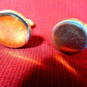 May include: Two oval-shaped cufflinks with a silver-colored border and a copper-colored center. The cufflinks are set against a red fabric background. The cufflinks have a small, yellow-colored post.