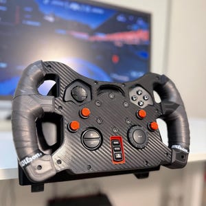 May include: A black racing steering wheel controller with a carbon fibre texture finish. It features grey grip handles with the Wilson logo, multiple circular orange buttons, a directional pad, action buttons, and a central PlayStation logo button.