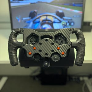 May include: A black carbon fiber racing steering wheel controller featuring a textured grip, a central D-pad, four PlayStation-style action buttons, four orange circular buttons, and a central dial, positioned in front of a computer monitor.