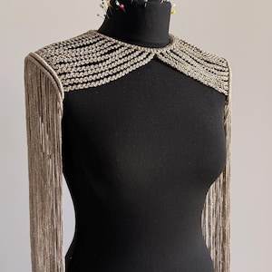 Peut inclure: Un accessoire d'&eacute;paule argent&eacute; m&eacute;tallique pr&eacute;sentant une s&eacute;rie de rang&eacute;es tress&eacute;es horizontales sur la zone de la clavicule et de longues franges fluides descendant des &eacute;paules. La pi&egrave;ce est pr&eacute;sent&eacute;e sur un mannequin noir.