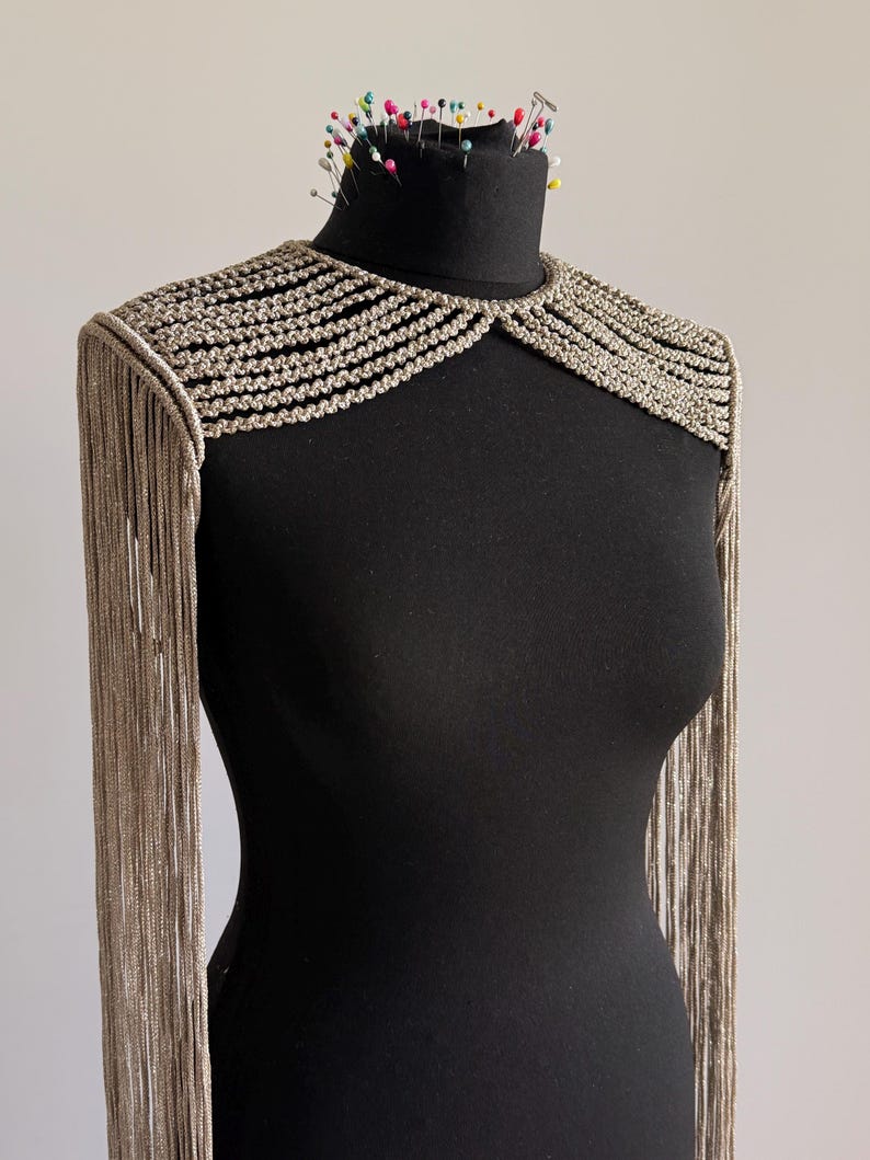 Peut inclure: Un accessoire d'&eacute;paule argent&eacute; m&eacute;tallique pr&eacute;sentant une s&eacute;rie de rang&eacute;es tress&eacute;es horizontales sur la zone de la clavicule et de longues franges fluides descendant des &eacute;paules. La pi&egrave;ce est pr&eacute;sent&eacute;e sur un mannequin noir.
