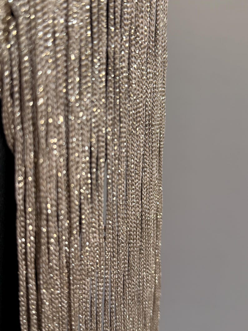 Peut inclure: Un agencement vertical de brins de franges scintillants couleur champagne. Les fils individuels sont &eacute;troitement tiss&eacute;s avec une finition m&eacute;tallique, cr&eacute;ant un rideau dense et textur&eacute; de mati&egrave;re r&eacute;fl&eacute;chissant la lumi&egrave;re en lignes droites.