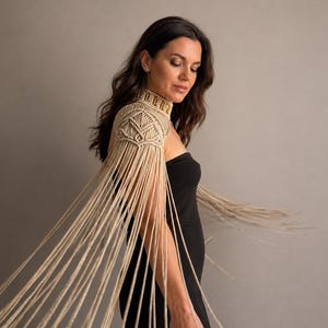 May include: A beige macrame shoulder piece featuring an intricate knotted collar and structured shoulder detailing, with long, flowing fringe strands extending downwards from the shoulder area, creating a dynamic and textured appearance.