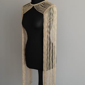 Peut inclure: Une pi&egrave;ce d'&eacute;paule en macram&eacute; beige avec un col montant et des d&eacute;tails nou&eacute;s complexes autour du cou. De longs fils de franges fins et pendants s'&eacute;tendent des &eacute;paules jusqu'&agrave; la taille, cr&eacute;ant un aspect textur&eacute; et superpos&eacute;.